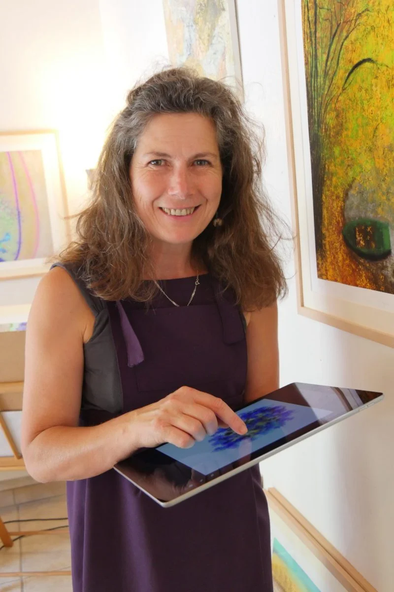 Anne smiling at us while holding a tablet with her finger on the screen showing a colorfull image, standing in front of a passe partout in her gallery
